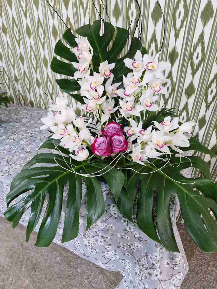 Arreglo floral con orquídeas blancas, rosas rosadas y grandes hojas verdes.