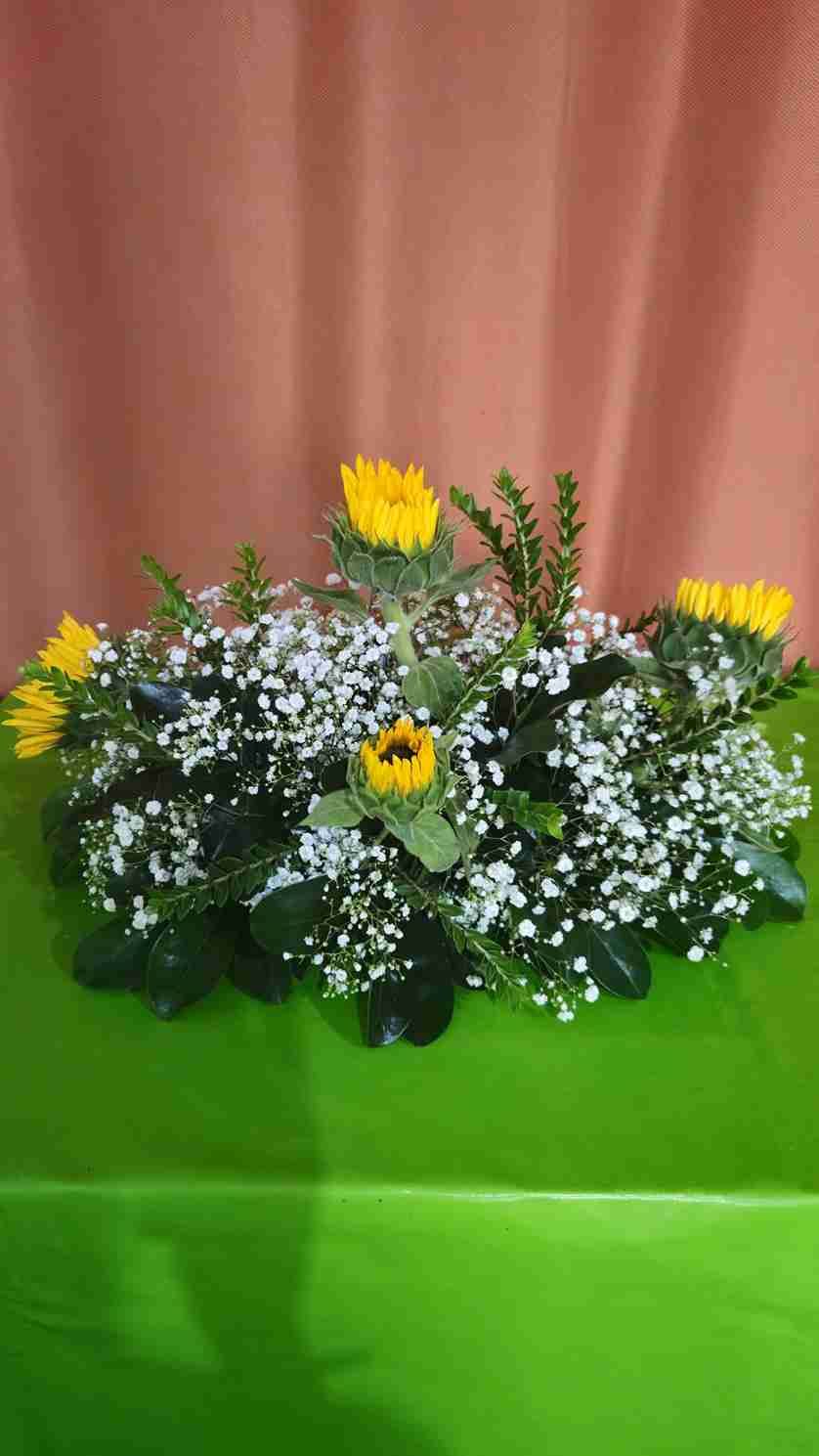 Arreglo floral con girasoles amarillos, gipsóforos blancos y follaje verde, sobre una superficie verde contra un fondo color durazno.