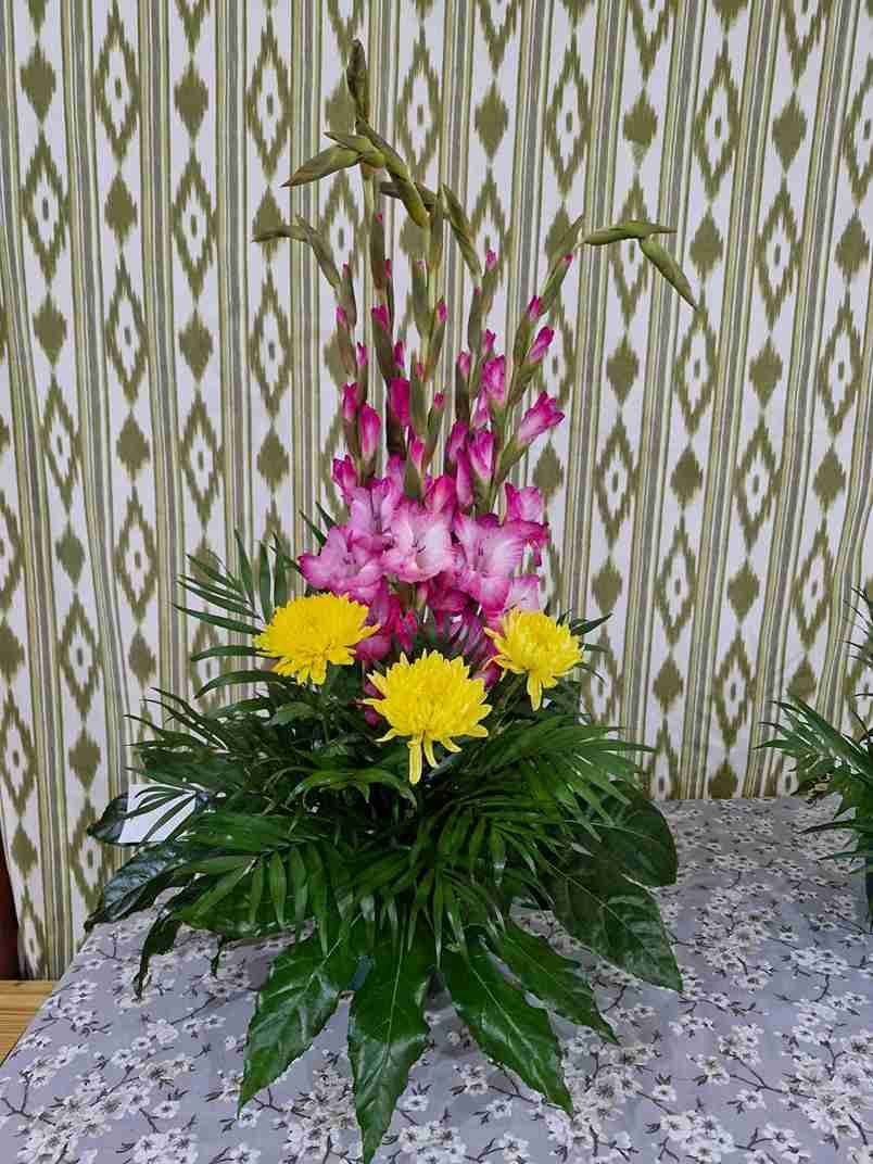 Arreglo floral: gladiolos rosados, crisantemos amarillos, follaje verde sobre un mantel estampado.