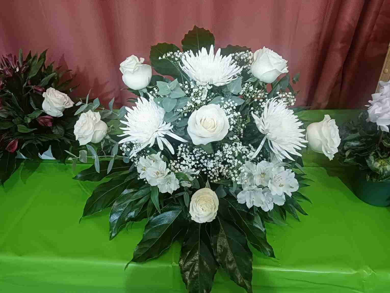 Arreglo floral blanco sobre una superficie verde, con rosas, crisantemos y follaje.