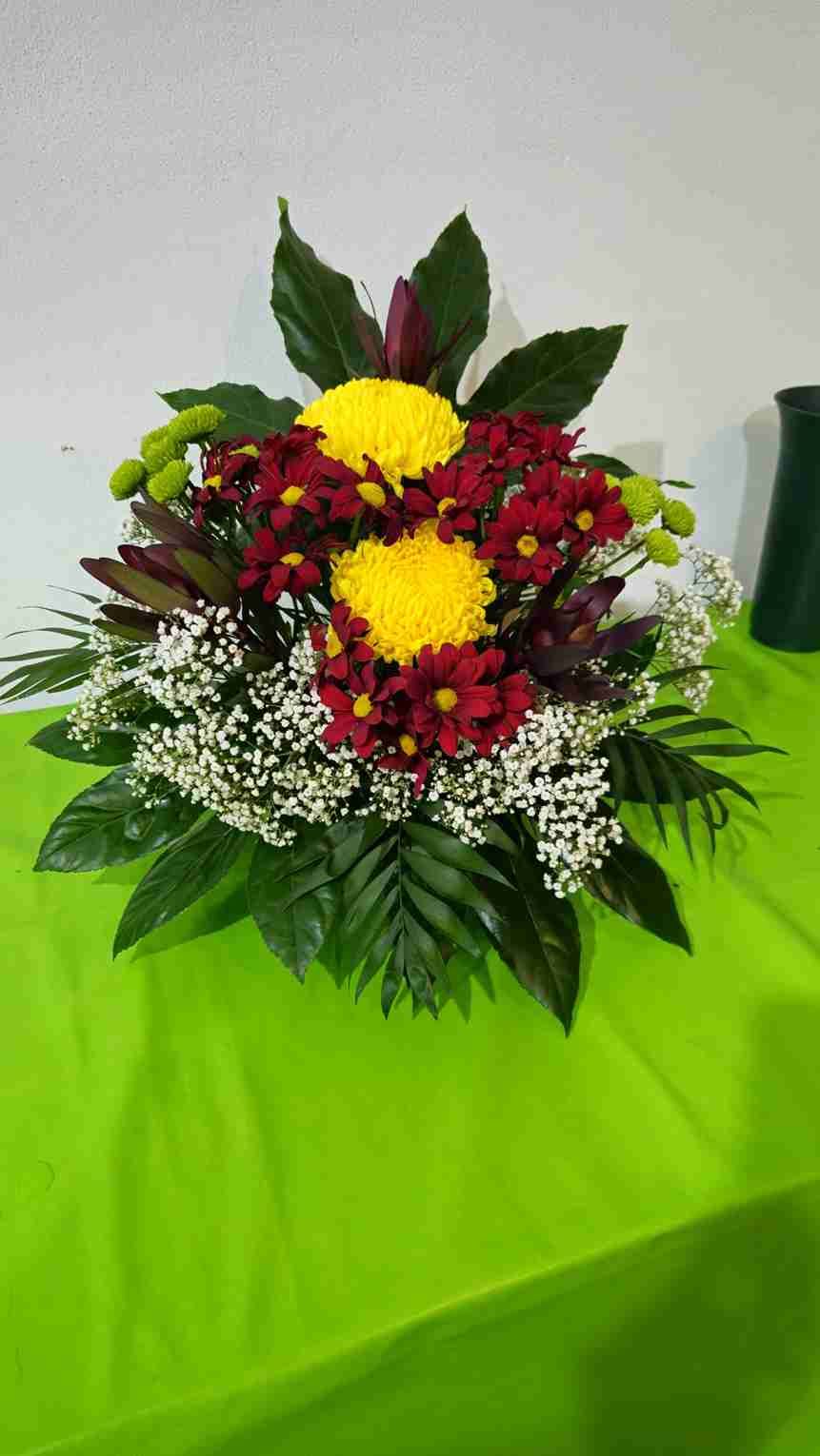 Arreglo floral sobre mantel verde: crisantemos amarillos y rojos con acentos verdes y blancos.