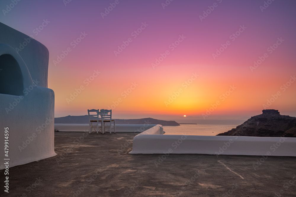 Zwei weisse Stühle auf einer Steinterrasse mit Blick auf das Meer bei Sonnenuntergang, der Himmel verläuft in sanften Farbverläufen von Lila zu Orange.