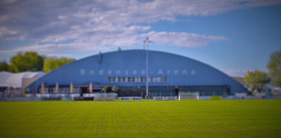 Hinter einem hellgrünen Feld unter einem teilweise bewölkten Himmel steht ein grosses, bogenförmiges, blaues Gebäude mit der Aufschrift „Bodensee-Arena“.