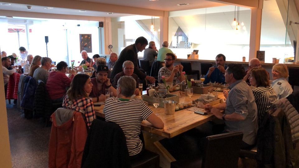 In einem hellen, modernen Restaurant sitzen die Menschen an einem langen Holztisch, essen und unterhalten sich bei einem geselligen Beisammensein.