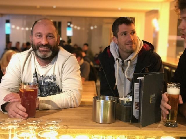 Zwei Personen sitzen an einer hölzernen Bar, halten Getränke in den Händen und blicken in die Kamera in einer warm beleuchteten Innenumgebung.