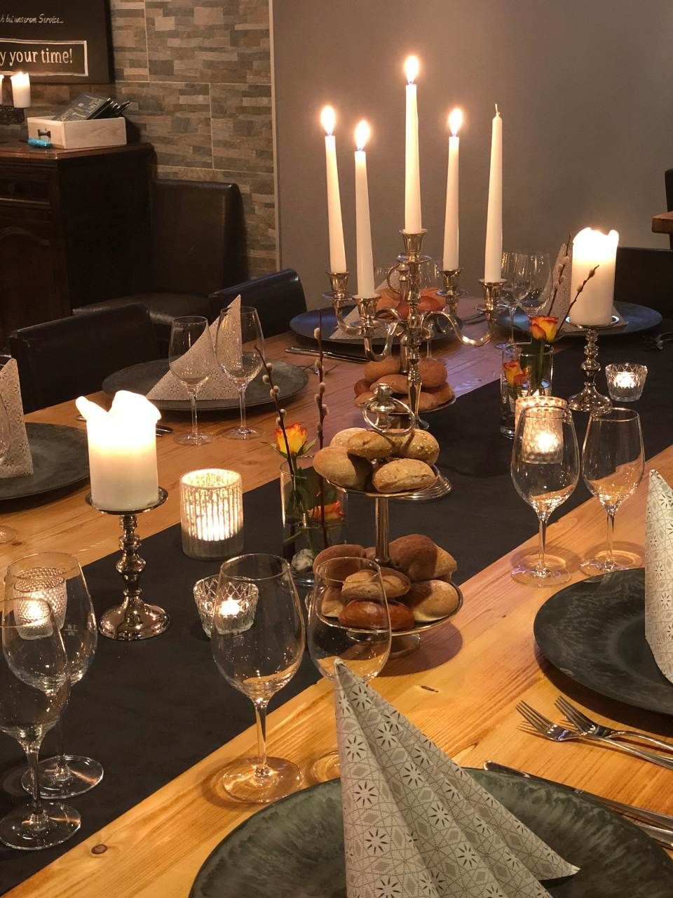 Ein formell gedeckter Esstisch mit Kerzen, Weingläsern, dunklen Servietten und einer Etagere mit Brötchen.