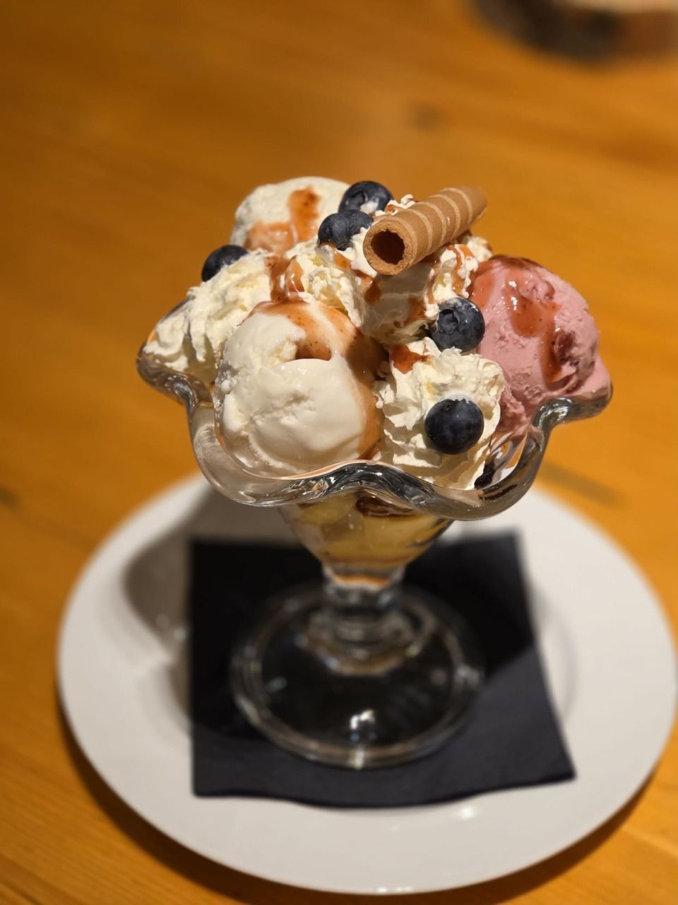 Ein Dessert-Eisbecher in einer Glasschale auf einem weissen Teller, bestehend aus Kugeln Eiscreme, Blaubeeren, Sirup und einer Waffelrolle.