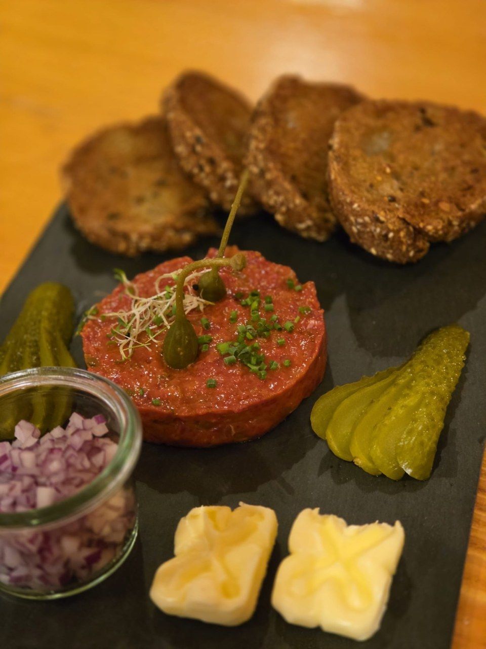 Ein Steak-Tartar-Gericht, serviert auf einem Schieferbrett mit Roggenbrot, Gurkenscheiben, gewürfelten Zwiebeln, Kapern und Butterstückchen.