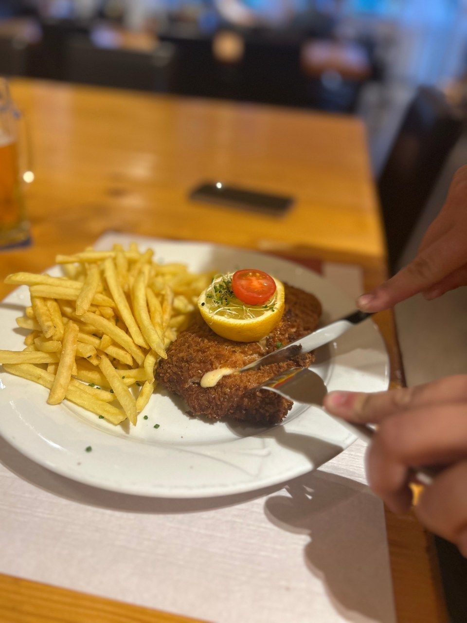 Ein Teller mit paniertem Schnitzel, Pommes frites und einer Zitronenscheibe, garniert mit einer Tomate, wird auf einem Tisch mit Messer und Gabel geschnitten.