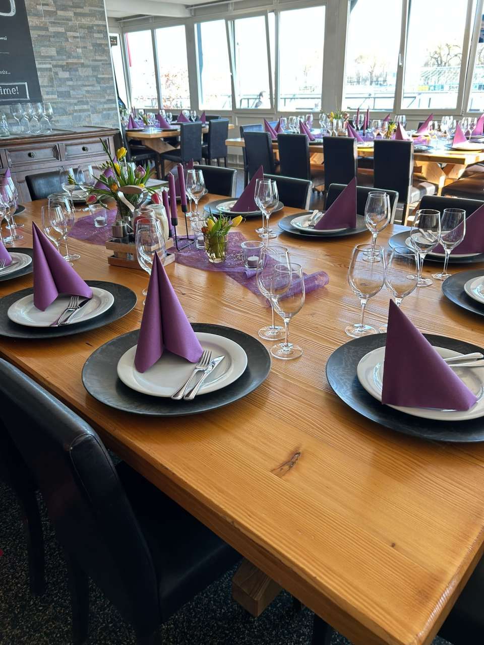 Ein Restauranttisch, gedeckt mit Tellern, Besteck und violetten Stoffservietten, im Hintergrund verschwommene Esstische.