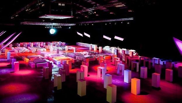 Ein abgedunkelter Veranstaltungsort, gefüllt mit Reihen kleiner, beleuchteter Stehtische und Sitzgelegenheiten, die in leuchtendes Neonlicht getaucht sind.