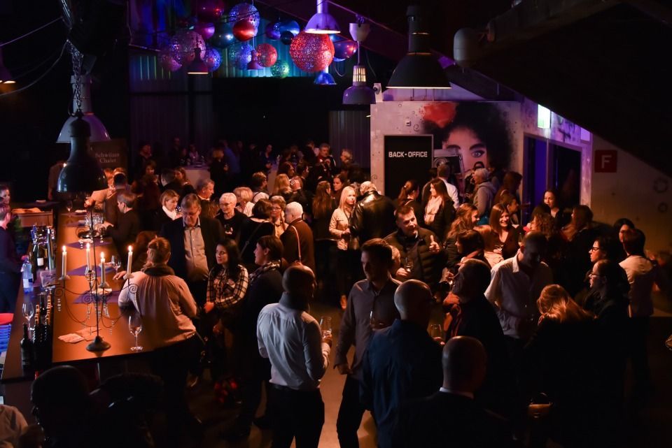 Eine überfüllte, schwach beleuchtete Bar im Innenbereich mit Pendelleuchten, Wandkunst und vielen Menschen, die sich unterhalten und miteinander plaudern.