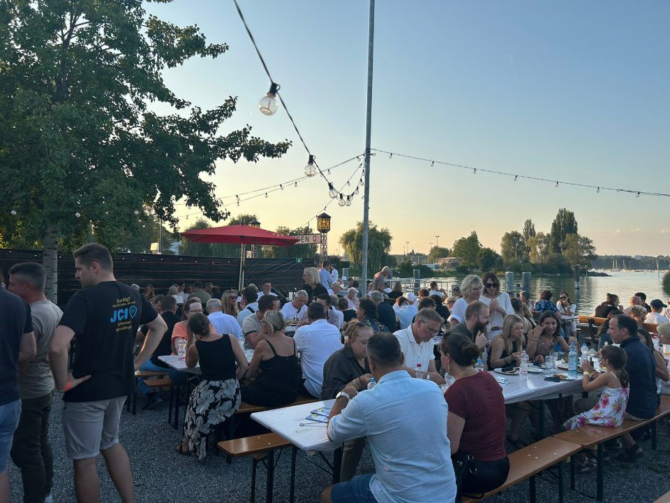 Bei Sonnenuntergang versammeln sich die Menschen an langen Tischen im Freien an einem See, der von Lichterketten beleuchtet wird, um gemeinsam zu essen.