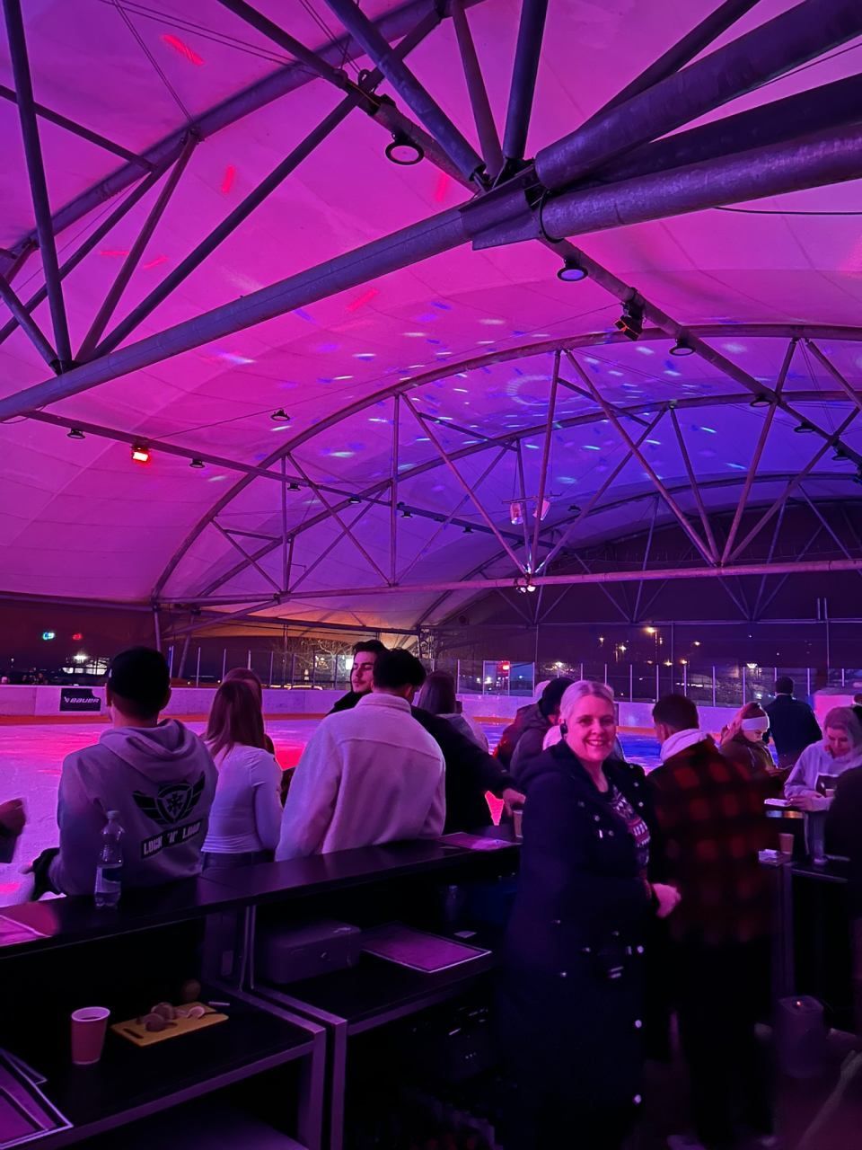 Menschen stehen an einem Bartresen und blicken auf eine beleuchtete Eislaufhalle, die in violettes und blaues Licht getaucht ist.
