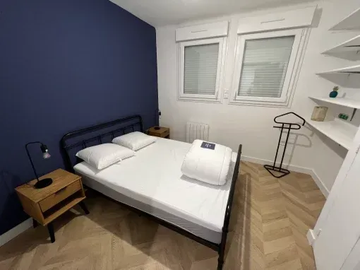 Chambre avec un mur bleu, un lit, deux fenêtres, un plancher en bois et une petite étagère.