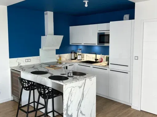 Cuisine blanche moderne avec comptoirs en marbre, murs bleus et tabourets de bar noirs.