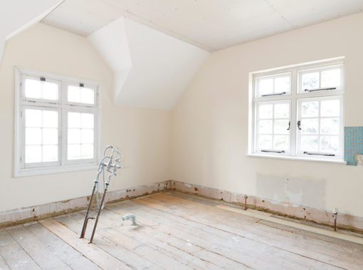 Chambre vide en cours de rénovation avec murs blancs, fenêtres et planchers apparents.