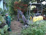 Personne en veste verte à côté d’une petite excavatrice en train de débroussailler un sentier de jardin
