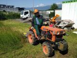 Un tracteur orange avec un ouvrier qui tond l’herbe au bord d’une route, avec un camion blanc en arrière-plan