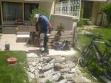 Personne accroupie dans un jardin, ramassant des morceaux de béton cassés près d’une terrasse et d’un vélo.