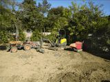 Engins de chantier sur un terrain en terre, près d’arbres et d’une zone boisée, avec un véhicule jaune et une remorque-benne rouge.