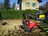Patio avec des chaises longues rouges et noires près d’une haie, avec une personne à côté d’une chaise jaune et une maison en arrière-plan