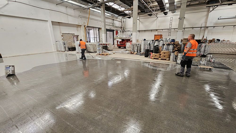 Arbeiter in orangefarbenen Westen begutachten einen glänzenden, nassen Betonboden in einem Lagerhaus.