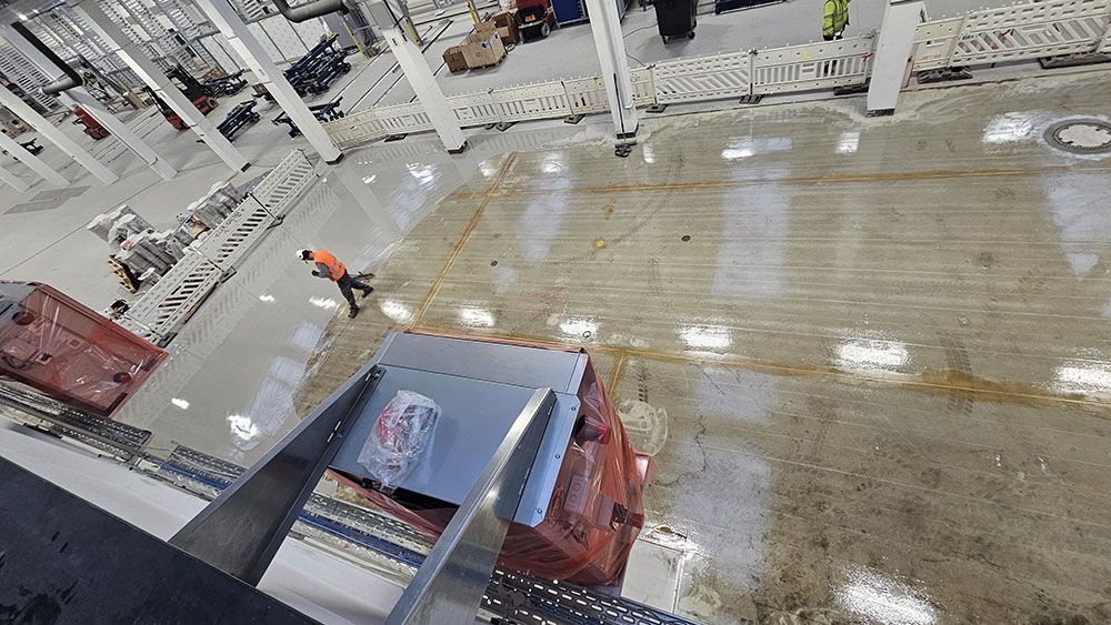 Ein Bauarbeiter in orangefarbener Weste geht über nassen Betonboden. Industrieumgebung mit Baumaschinen.