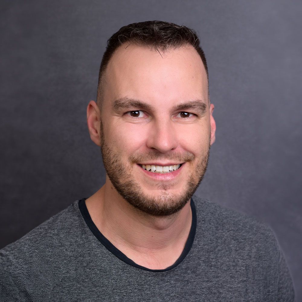 Ein Mann mit kurzem braunem Haar und Bart lächelt vor einem grauen Hintergrund. Er trägt ein graues Hemd.