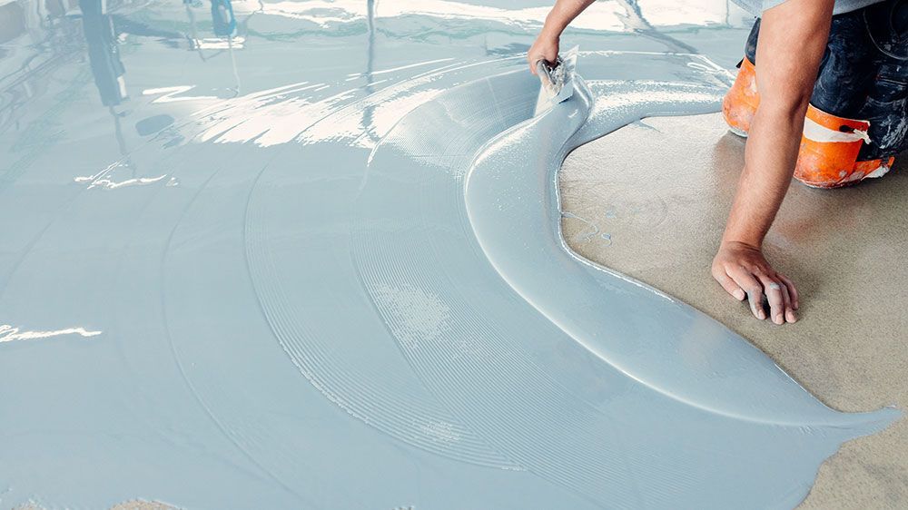 Eine Person glättet mit einer Kelle eine graue, flüssige Bodenbeschichtung auf einer Betonfläche.