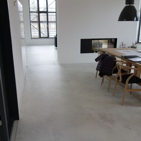 Großzügiges, modernes Zimmer mit hellgrauem Boden, Kamin, Esstisch und großen Fenstern.