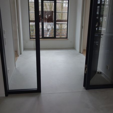 Leerer Raum mit hellgrauem Boden, großem Fenster und offenen, schwarz gerahmten Türen.