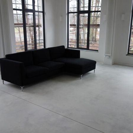 Schwarzes Ecksofa in einem hellen, weiß getünchten Zimmer mit großen Fenstern und Blick auf Bäume.