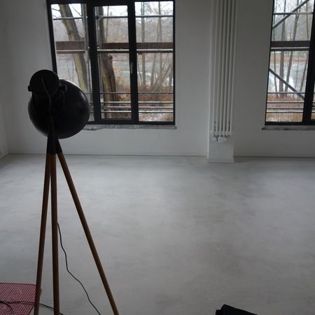 Leeres Zimmer mit hölzerner Dreibeinlampe. Große Fenster bieten Ausblick nach draußen. Heller Boden und helle Wände.