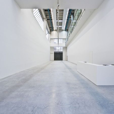 Langer, weißer Galerieraum mit Betonboden. Große schwarze Tür am Ende, natürliches Licht von oben.