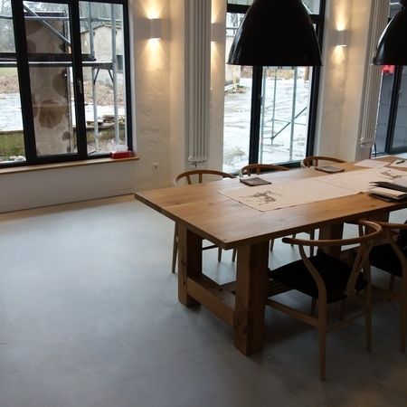 Ein großer Esstisch aus Holz mit Stühlen in einem Zimmer mit großen Fenstern und hellen Wänden.