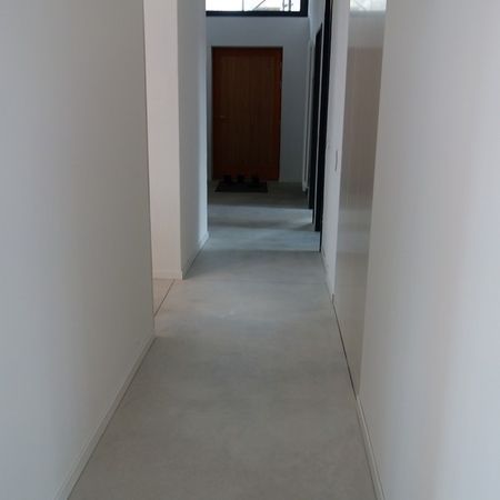 Schmaler Flur mit weißen Wänden und grauem Boden. Am Ende befindet sich eine braune Tür.