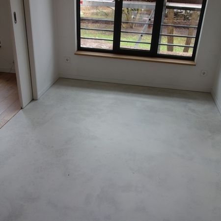 Leerer Raum mit hellgrauem Betonboden und Fenster mit schwarzem Rahmen.