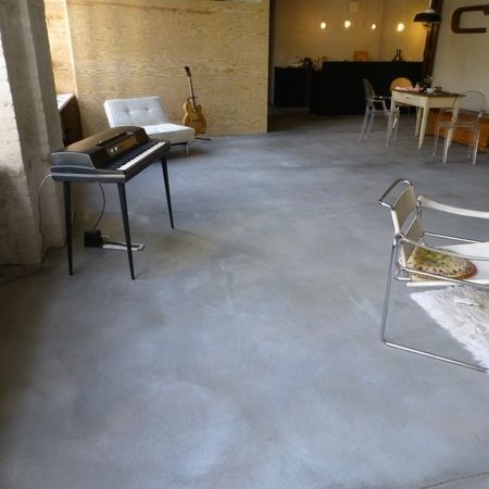 Zimmer mit Betonboden, Klavier, Gitarre, Stühlen und Tisch. Natürliches Licht fällt durch ein Fenster.