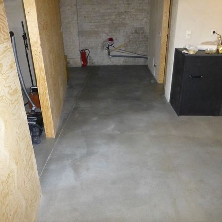 Betonboden in einem Raum mit Sperrholzverkleidung und schwarzem Schrank; eine Tür führt zu einem Hauswirtschaftsraum.