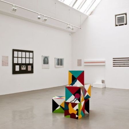 Galerieraum mit farbenfrohen, geometrischen Skulpturen und verschiedenen gerahmten Kunstwerken an weißen Wänden.