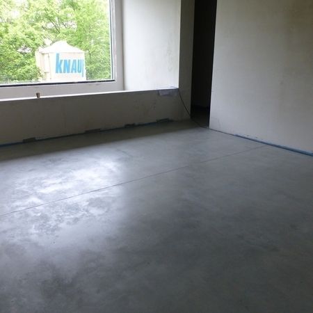 Ein leerer Raum mit glattem, grauem Betonboden. Durch ein Fenster ist ein Teil des Gebäudes mit dem Logo „KNAUF“ zu sehen.