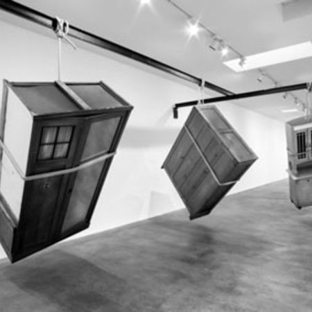 Drei hölzerne Gebilde, die Schränken ähneln, hängen in einem Galerieraum schräg von der Decke.