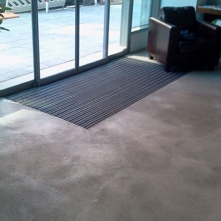 Eine rechteckige, schwarze Metallgittermatte vor Glastüren im Inneren eines Gebäudes mit einem Stuhl.