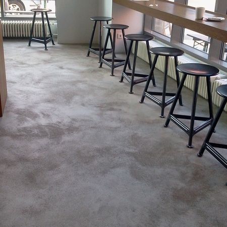 Grauer Betonboden mit schwarzen Barhockern in einem Café-Ambiente, neben einer langen Theke und Fenstern.
