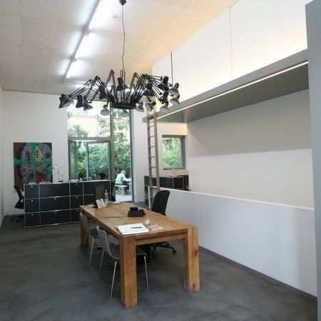 Modernes Büro mit Holztisch, dunklen Schränken, einzigartigem Kronleuchter und einer Person an einem Schreibtisch am Fenster.