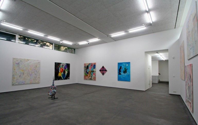 Interieur einer Kunstgalerie mit abstrakten Gemälden an weißen Wänden, Neonröhren und dunklem Boden.