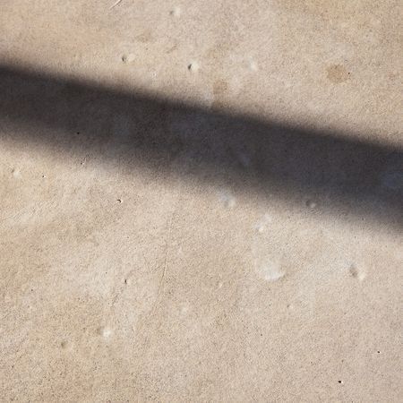 Schatten eines Objekts, der diagonal über eine strukturierte beige Oberfläche mit kleinen dunkleren Flecken fällt.