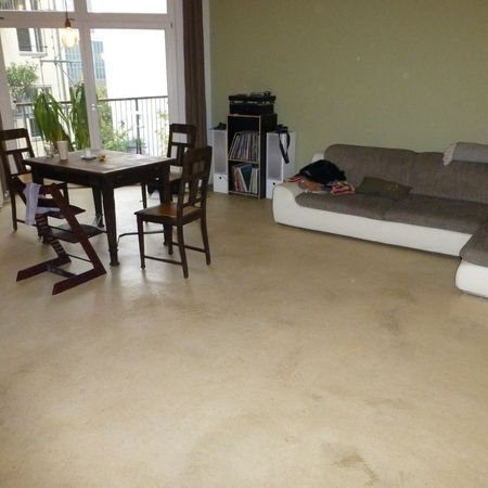 Großzügiges Wohnzimmer mit Esstisch, Ecksofa und Balkon mit Fenster, hellen Bodenbelägen und salbeigrünen Wänden.