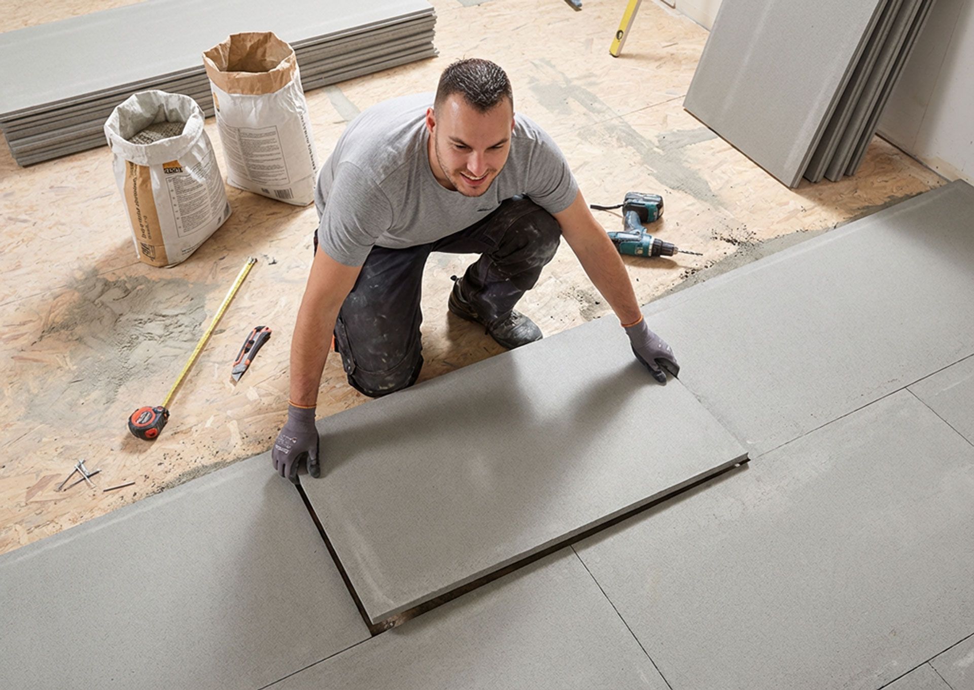 Ein Mann verlegt kniend lächelnd graue Bodenfliesen; im Raum befinden sich Säcke mit Zement, Werkzeug und gestapelte Fliesen.